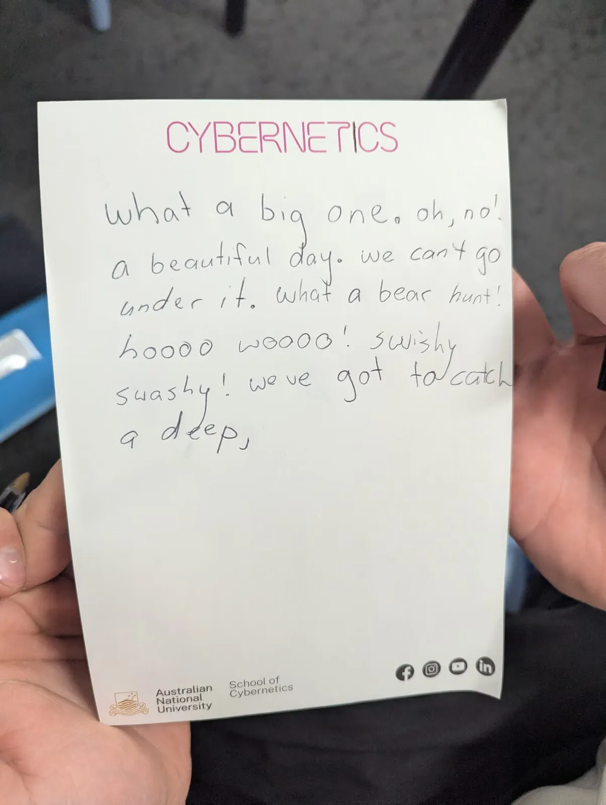 A student holding up their handwritten workshop notes, headed "Cybernetics"