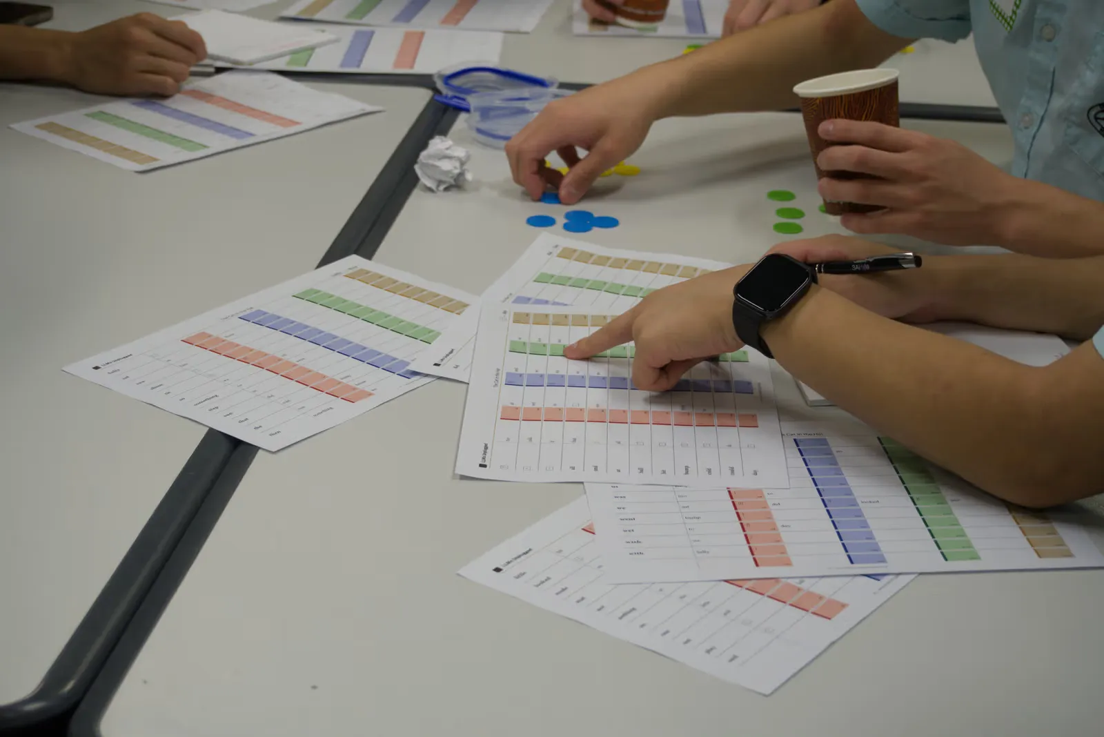 Students' hands on a table of colour-banded ledger sheets, one pointing at a row while another holds a paper cup beside a scatter of blue, green and yellow counters