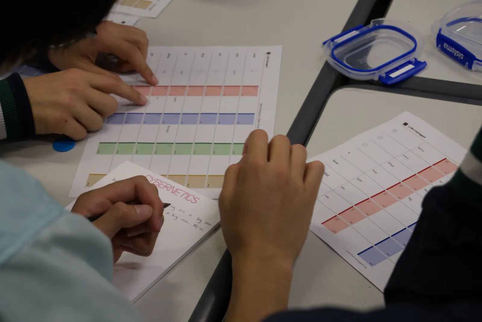 Two students' hands tracing rows of a ledger sheet, one holding a pen over a notepad of generated words