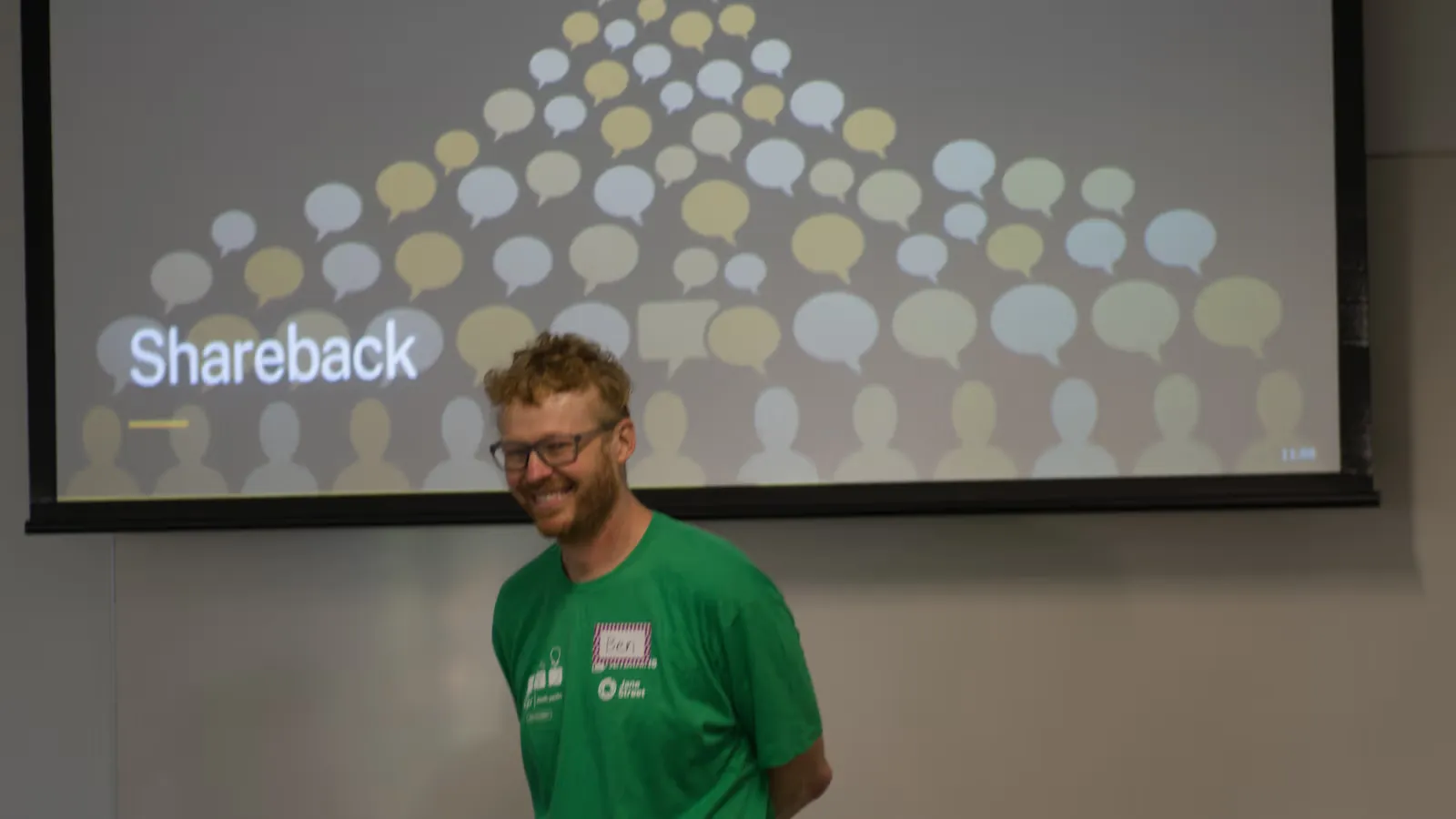 Ben, in a green t-shirt, grinning in front of a slide headed "Shareback" that shows a crowd of speech bubbles above simple silhouettes